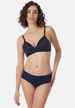 Nainen, jolla on kiharat tummat hiukset, mallintaa yhteensopivaa laivastonsinistä rintaliivi- ja alusvaatesettiä yksiväristä valkoista taustaa vasten.