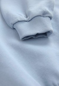 Tessuto di felpa blu chiaro che mostra un polsino a coste e una texture liscia, con messa a fuoco ravvicinata sui dettagli del materiale e della cucitura.