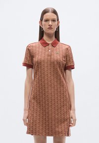 Pólóruha geometrikus barna mintával, piros gallérral és mandzsettákkal, oldalsó hasítékokkal és kis zöld logóval a mellrészen.