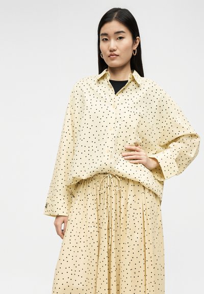 Νέα γυναίκα φοράει μπεζ πουκάμισο με κουμπιά και παντελόνι φαρδύ με μαύρες πουά, στέκεται με το ένα χέρι στη μέση.