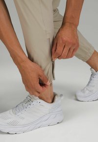 Beige träningsbyxor med dragkedjor vid bensluten, lättviktsmaterial och en slät textur. Bärs med vita löparskor med meshdetaljer.