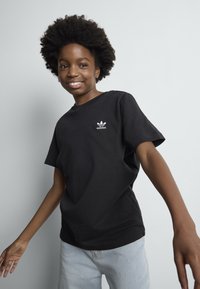Smilende person med afrofrisyre iført svart Adidas t-skjorte og lyseblå jeans, poserer mot en ensfarget lys grå bakgrunn.