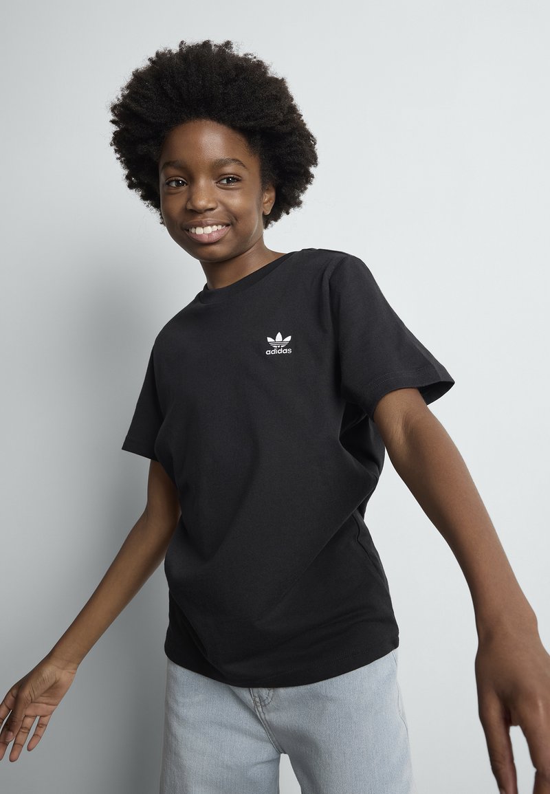 Smilende person med afrofrisyre iført svart Adidas t-skjorte og lyseblå jeans, poserer mot en ensfarget lys grå bakgrunn.