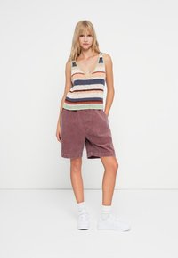 Gestreiftes Strick-Top in Pastellfarben, kombiniert mit bordeauxfarbenen Corduroy-Shorts. Das Model trägt weiße Sneakers und Socken. Einfaches, lässiges Outfit.