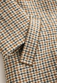 Beidzis un brūns houndstooth raksta audums ar mīkstu tekstūru, kas attēlo tuvplānu skatu uz šuvēm salocīta apģērba piedurknei.