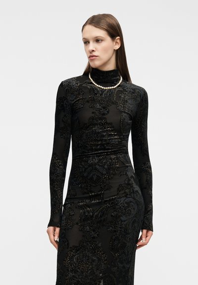 Γυναίκα που φοράει εφαρμοστό μακρυμάνικο μαύρο φόρεμα με βελούδινα λουλουδένια σχέδια και περιδέραιο από πέρλες, κοιτάζοντας ελαφρώς προς τα αριστερά της.