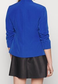 Blå skuren blazer med rynkade ärmar, strukturerad design och slät textur, i kombination med en svart läderkjol med utsvängd form.