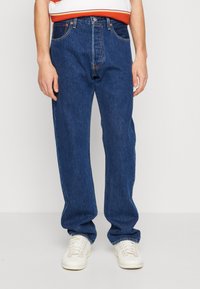 Vaqueros de mezclilla azul oscuro con corte recto, cinco bolsillos y detalles de costura sutiles; combinados con zapatillas blancas.