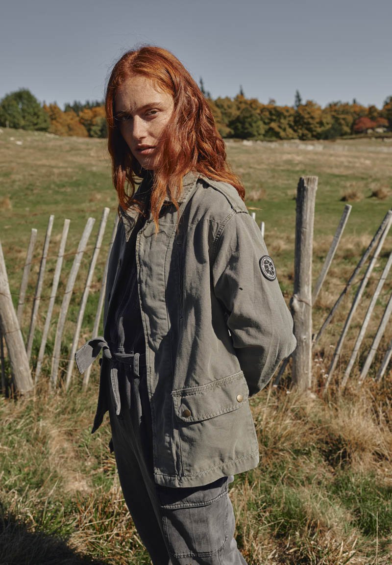 Olijfgroene, taille-lange jas met een capuchon, ontworpen met een strik taille, voorzakken en een logo-embleem op de mouw. Natuurlijke buitenomgeving.