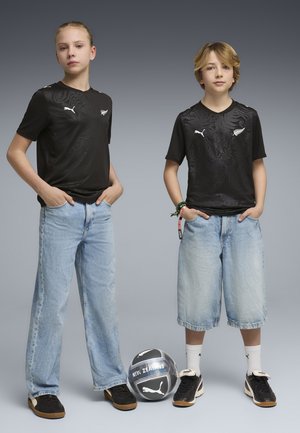Dwoje dzieci ubranych w czarne koszulki Puma Nowa Zelandia oraz dżinsy stoi na szarym tle z piłką do piłki nożnej Puma Nowa Zelandia.