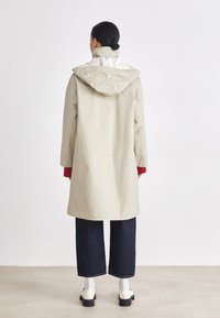 Beige lightweight coat with a hood, featuring a high collar and contrasting white lining. Paired with dark jeans and cream shoes.