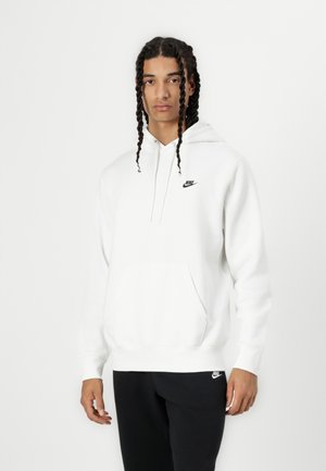Sudadera blanca de Nike con un bolsillo frontal, capucha con cordón y pequeño logotipo negro en el pecho. Combinada con pantalones negros. Textura suave.