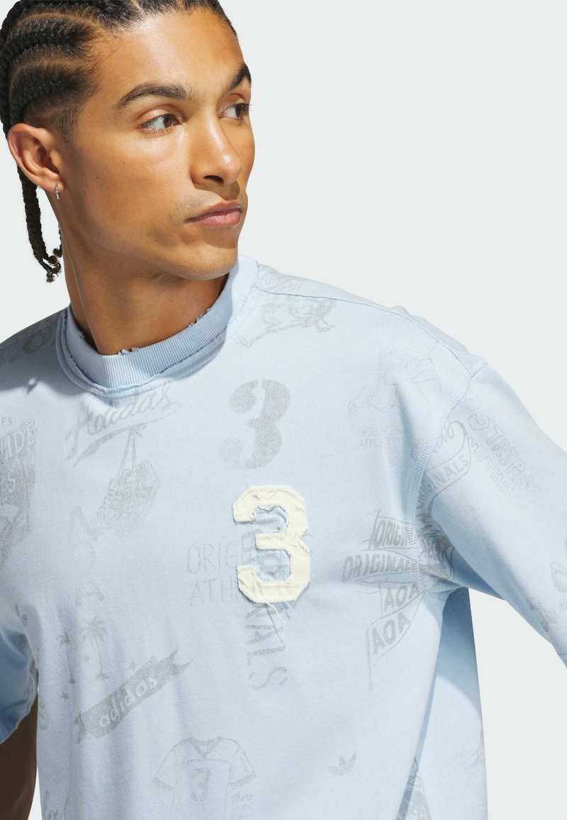 Hombre con cabello trenzado que lleva una sudadera azul claro con un gran número 3 texturizado y estampados gráficos descoloridos, mirando hacia su izquierda.