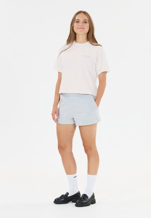 Jeune femme debout portant un t-shirt blanc, un short gris clair, des chaussettes blanches et des mocassins noirs, sur un fond blanc uni.