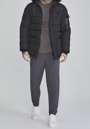 Homme portant une veste matelassée noire avec capuche en fourrure, pantalon gris, chemise grise, baskets blanches, et tatouages visibles sur les mains et le cou.