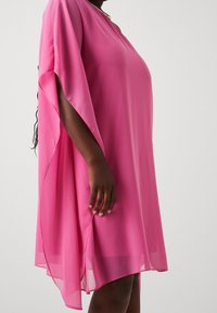 Personne à la peau foncée portant une robe courte, ample et rose vif à manches courtes, avec un ourlet asymétrique flottant, se tenant devant un fond blanc.