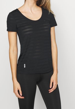 Vrouw die een nauwsluitende zwarte top met korte mouwen draagt met subtiele horizontale strepen en nauwsluitende zwarte legging tegen een effen lichte achtergrond.