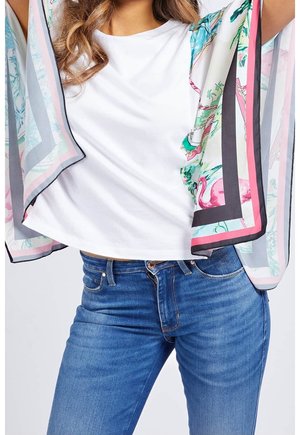 Kvinde iført blå jeans og hvid t-shirt, der holder et farverigt gennemsigtigt tørklæde med pink, grønne og sorte mønstre omkring skuldrene.