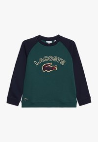 Felpa verde con maniche raglan blu navy, dotata di un logo Lacoste ricamato e un design di coccodrillo sul davanti. Tessuto morbido.