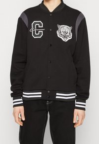 Svart varsityjacka med kontrasterande grå axelpaneler, vit broderad tiger grafik, samt randiga ribbade cuffar och nederkant. Framknäppning med tryckknappar.