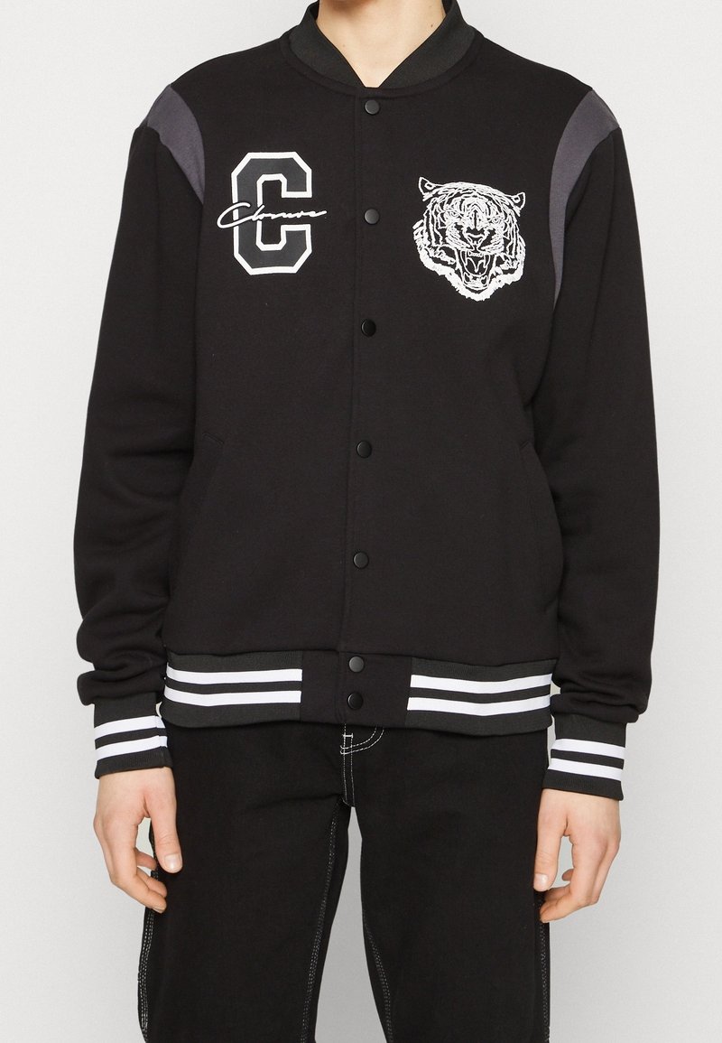 Svart varsityjacka med kontrasterande grå axelpaneler, vit broderad tiger grafik, samt randiga ribbade cuffar och nederkant. Framknäppning med tryckknappar.