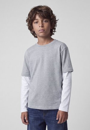 Garçon aux cheveux bruns bouclés portant un t-shirt gris à manches courtes par-dessus un t-shirt blanc à manches longues et un jean bleu, debout devant un fond uni.