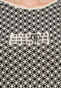 Črno in kremno vzorčasta pletena jopica s geometrijskim vzorcem in vdelanim logotipom na sredini. Okrogla izrezana ovratnica, teksturirana tkanina.