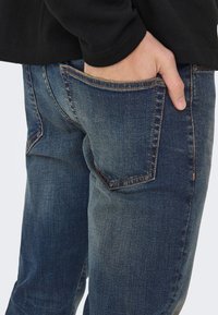 Tumši zili džinsa džinsi ar šauru piegriezumu, ar oranžiem šuvumiem un priekšējo kabatu. Audumam ir viegli izbalējusi tekstūra.