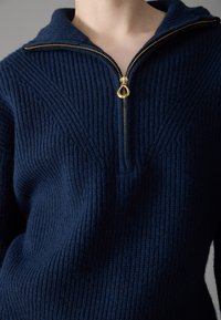 Maglione blu navy con zip, caratterizzato da una texture a coste, con colletto alto e zip dorata. Dettaglio di cucitura a forma di V visibile sul davanti.