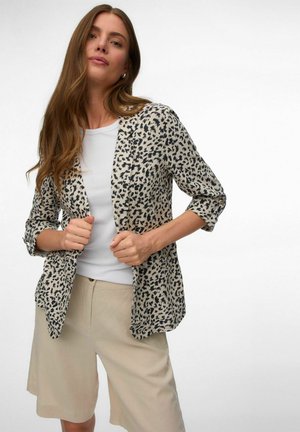 Femeie cu păr lung, castaniu, purtând un top alb, pantaloni scurți bej și un blazer bej cu model leopard, cu mânecile suflecate.