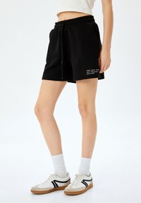 Pantalones cortos de algodón negros con cinturilla elástica y cordón, que presentan el texto en blanco "abre tu mente y descúbrete a ti mismo" en el lateral.