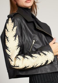 Vrouw draagt een zwarte leren jas met beige bladeren in bladvorm en gouden studs langs de mouwen, terwijl ze de voorkant van de jas vasthoudt.