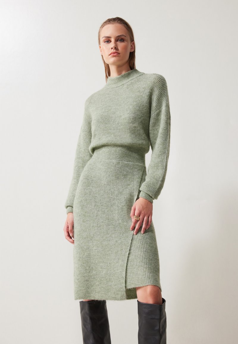 Robe en tricot de couleur vert clair, douce et légère, avec un col montant et des manches longues. Possède une fente sur le côté et une texture côtelée. Associée à des bottes noires hautes.