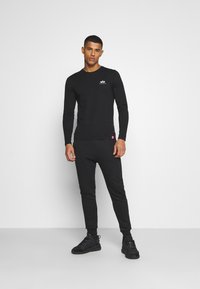 Schwarzes Langarmshirt mit Rundhalsausschnitt und Logo, kombiniert mit passenden schwarzen schmal geschnittenen Hosen und schwarzen Sneakers. Glatter Stoff, figurnahes Design.