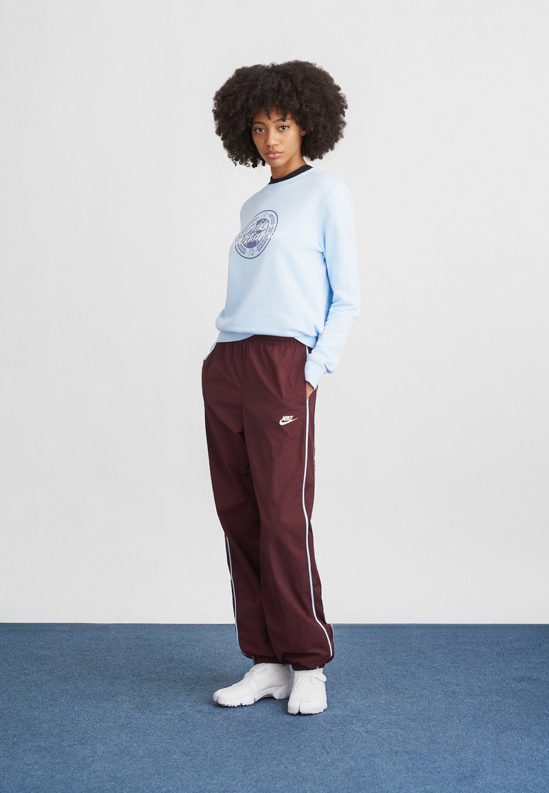 Sweatshirt bleu clair avec un logo rond, associé à un pantalon de survêtement bordeaux avec des rayures latérales, complété par des baskets blanches sportives.