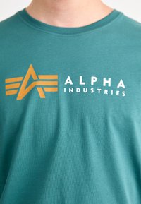 Camiseta de algodón color verde azulado que presenta un logo dorado con una 'A' y dos franjas horizontales, junto con 'ALPHA INDUSTRIES' en texto blanco.