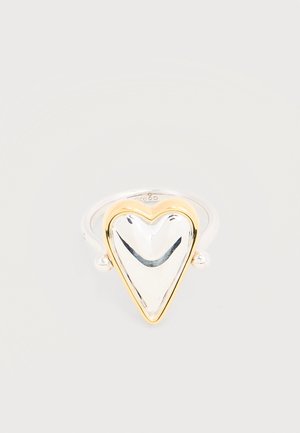 Srebrny pierścionek z wypolerowanym serduszkowatym wierzchołkiem obwiedzionym złotem, prezentowany na gładkim jasnym tle.