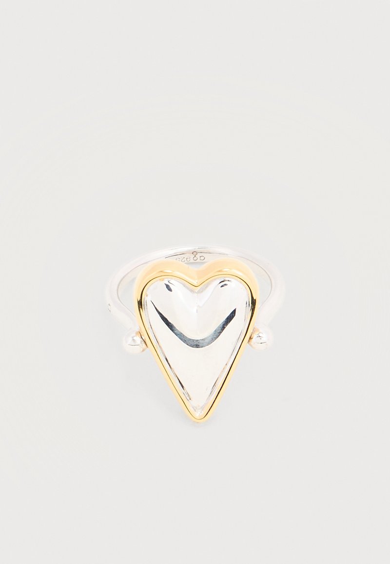 Ασημένιο δαχτυλίδι με λείο ανώτερο μέρος σε σχήμα καρδιάς, περιγραμμένο με χρυσό, απεικονιζόμενο σε απλό φωτεινό φόντο.