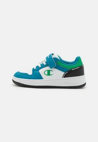 Der Schuh verfügt über ein Obermaterial aus weißem Leder mit blauen und grünen Akzenten, einem Klettverschluss, Perforationen zur Belüftung und einer schwarzen Gummisohle.