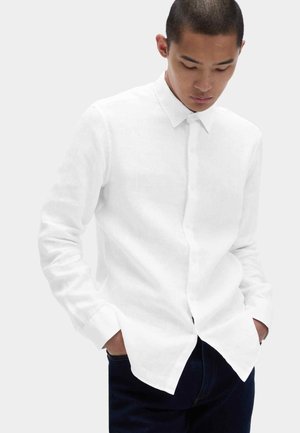 Junger Mann mit den Händen in den Taschen, trägt ein weißes, langärmeliges Hemd mit Knöpfen und dunkle Hose, schaut nach unten vor einem schlichten Hintergrund.