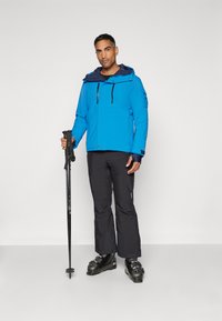 Helle blaue wasserdichte Jacke mit verstellbarer Kapuze, schwarzen Akzenten und Reißverschluss-Taschen. Schwarze Skihosen und -stiefel mit Skistöcken.