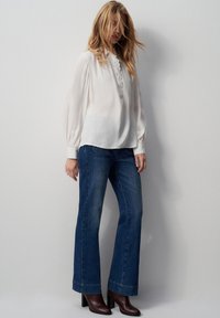 Blusa bianca a maniche lunghe con patta a bottoni, abbinata a jeans a zampa d'elefante blu scuro e stivali marroni con tacco, design minimalista.