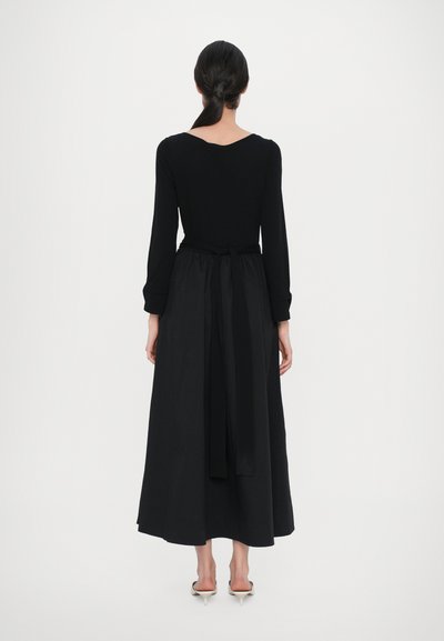 Ilga juoda suknelė su ilgomis rankovėmis, apvaliu iškirpimu ir surištu juosmeniu. Lygus audinys, laisvai krentanti sijono dalis siekia žemiau čiurnų.