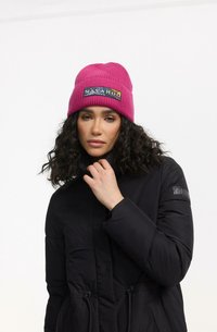 Roosa kudumine beanie, millel on sooniline tekstuur ja kangast silt, millel on kiri "NAVAHOO". Kombineeritud musta puffer-jacketiga, millel on tõmblukud.