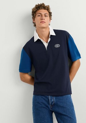Jeune homme aux cheveux bouclés portant un maillot de rugby à manches courtes bleu marine et bleu ainsi qu'un jean bleu, debout contre un mur blanc.