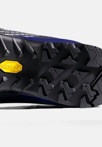 Suola Vibram con battistrada aggressivo, caratterizzata da una superficie nera testurizzata e dettaglio del logo giallo. Progettata per garantire trazione su diversi tipi di terreno.