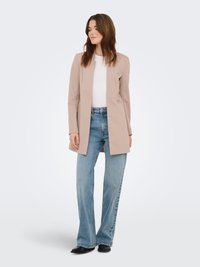 Veste beige à manches longues avec devant ouvert, associée à un t-shirt blanc et un jean évasé bleu clair, complétée par des chaussures noires.