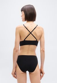 Γυναίκα που βλέπουμε από πίσω, φορά μαύρα εσώρουχα με σταυρωτούς ιμάντες στο σουτιέν, σε απλό ανοιχτό γκρι φόντο.