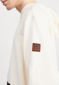 Vista lateral de una persona que lleva una sudadera blanca con un parche del logo de ellesse en naranja y negro en la parte superior de la manga.
