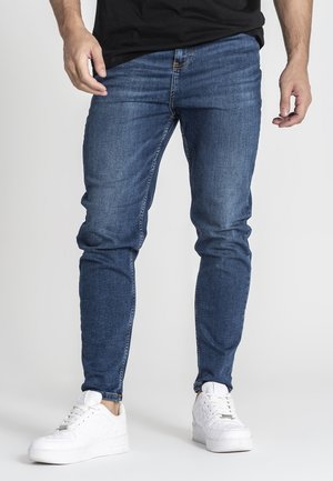 Uomo che indossa jeans blu slim fit e sneakers bianche in piedi davanti a uno sfondo chiaro e semplice.